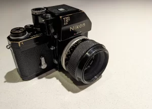 Nikon F with Photomic FTn. Camera is black with some brassing. The lens is also black with a silver aperture ring. It sits under bright light on a light coloured surface.